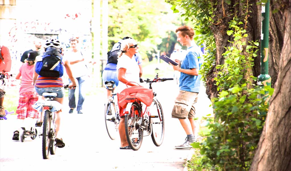 Ein Mann auf einem Fahrrad im freien spricht mit einem jungen Mann der ein Tablet in der Hand hält